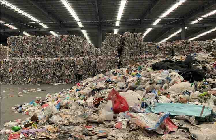 A large warehouse filled with huge stacks of compressed recyclable materials and scattered loose recyclables, creating a sense of industrial scale and environmental urgency.